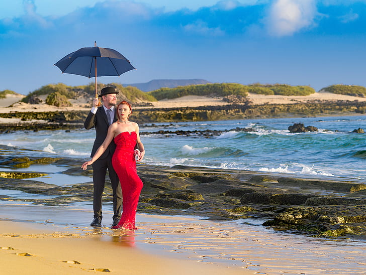 man in black suit holding black umbrella with woman in red dress on beach