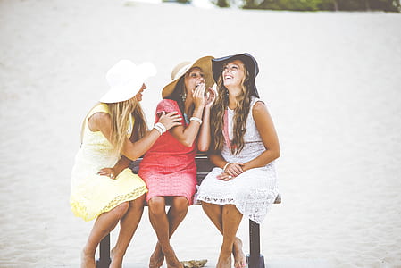 three women with assorted-color dresses sitting on bench beside seashore