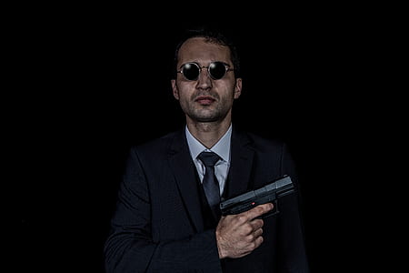 man in black suit jacket and black necktie holding black semi-automatic pistol with black hippie sunglasses