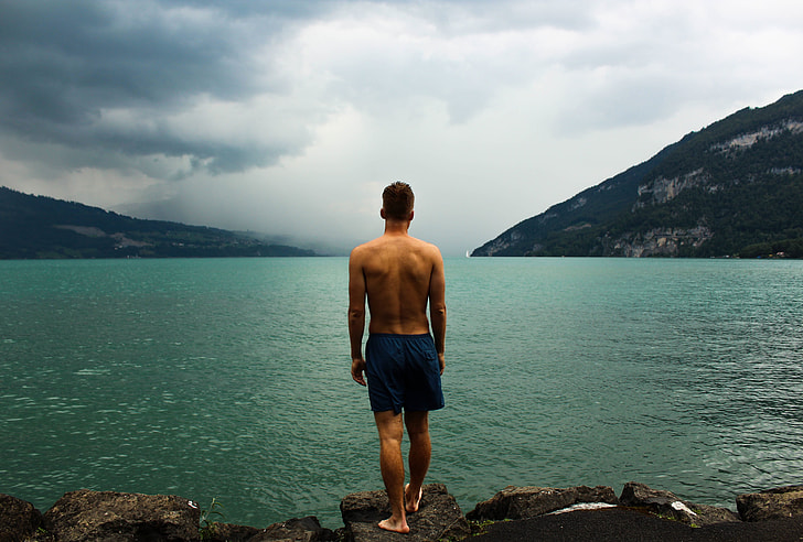 man in black short standing on rock beside lake