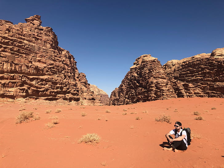 Man Wearing White Shirt and Black Pants Sitting on Soil Behind Rock Formation Mountain