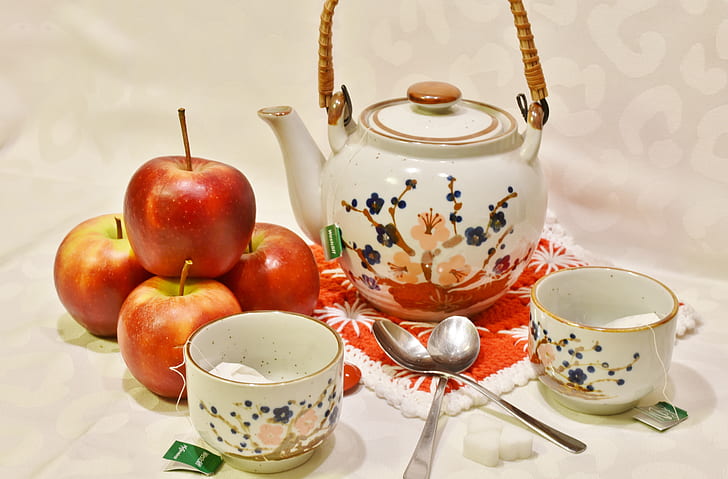 four red apples near white and multicolored ceramic teapot near two white ceramic teacups and two spoons