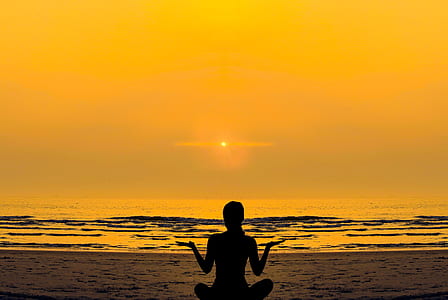 silhouette of woman meditating near body of water during sunset