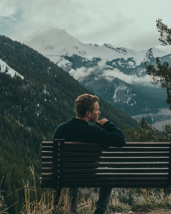 image contains man sitting on bench near mountains