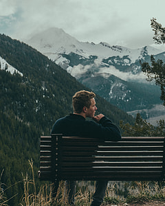 image contains man sitting on bench near mountains