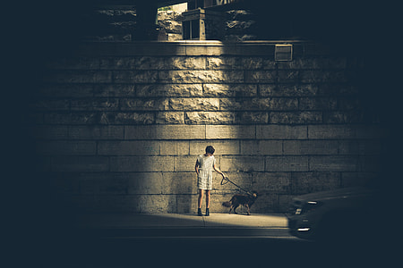 man in white shirt and white bottoms standing beside gray concrete wall while holding dog leash near car during nighttime