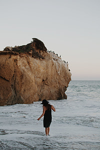 woman in black sleeveless bodycon mini dress and sun hat walking on seashore during daytime