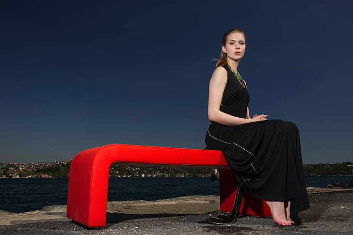 woman in black sleeveless dress sitting on red bench near body of water