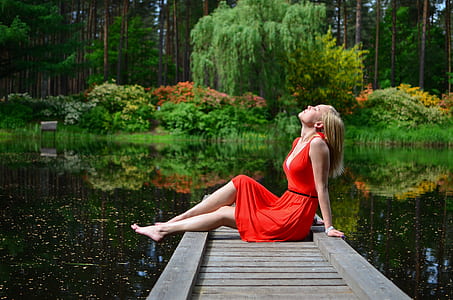photo of woman sitting near edge of dock surrounded by body of water far away from tall trees