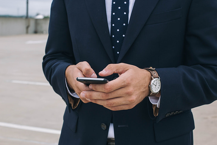 man in  blue suit jacket  and blue necktie holding smartphone during daytime