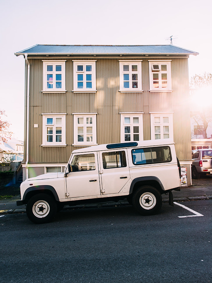 white Land Rover Defender SUV parked in front of brown 3-story building under gray clouds