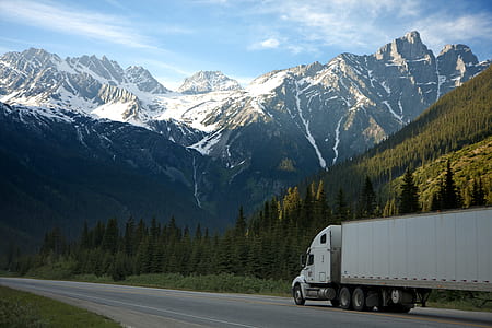 white freight truck running on concrete road near snow covered mountain at distance during daytime