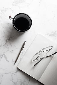 white and black ceramic mug beside notebook with pen and eyeglasses