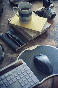 computer keyboard, mouse with mouse pad, mug, books and camera on brown wooden board