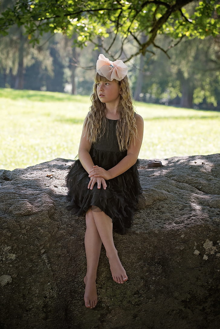 girl wearing black dress with pink bow accent headband sitting on grey rock under green leaf tree