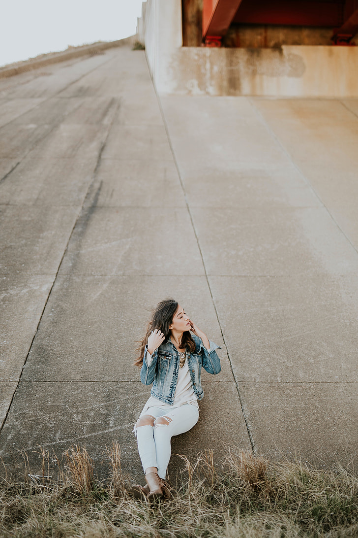 woman in blue washed denim jacket, white shirt, and distressed blue jeans leaning in concrete wall