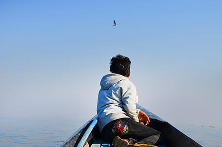 Man in Grey Hoodie and Black Pants Sitting in the Middle of Boat Looking at Bird in the Sky