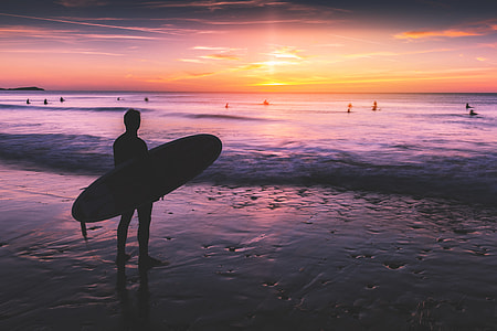 A surfer stands on the beach and looks out to the ocean at sunset. Image captured in Cornwall, England