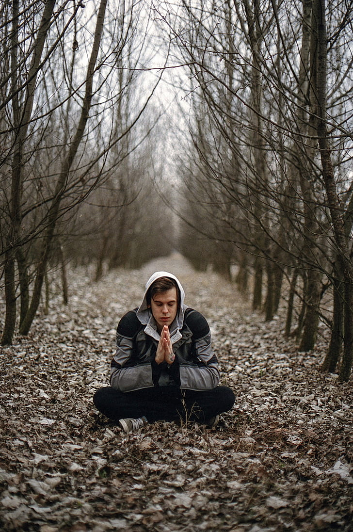 man wearing gray jacket and black pants meditating on road with dried leaves near trees
