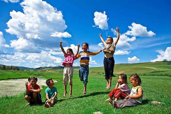 four children sitting on green grass and three children jumping taken under white clouds during daytime
