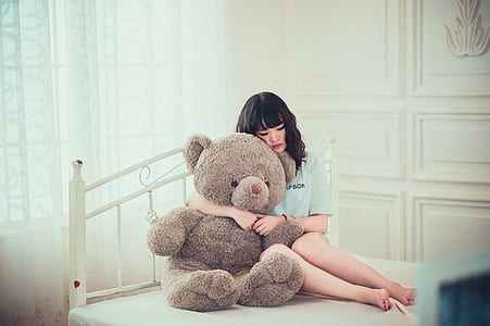 woman in white top hugging brown bear plush toy on bed
