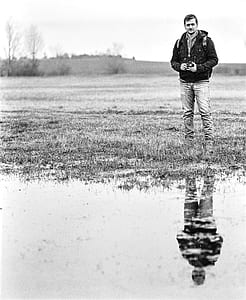 Man in Jacket Standing Near Body of Water Holding Camera in Grayscale Photography