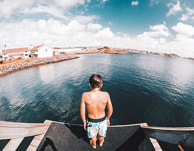 boy wearing blue shorts near body of water under white clouds during daytime