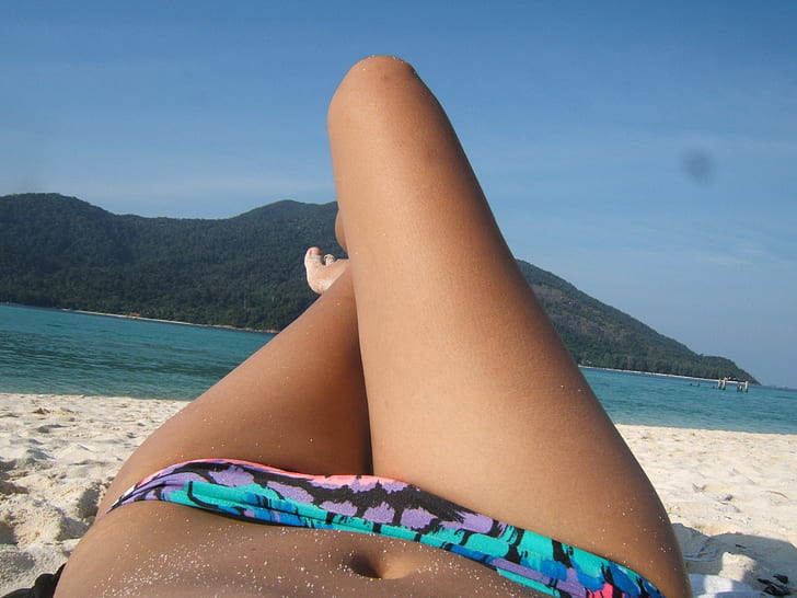 woman wearing blue and multicolored bikini bottom lying on white sand near blue sea at daytime