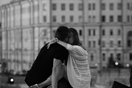 grayscale photography of woman kissing man near white building