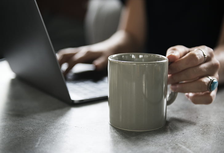 person using laptop computer about to reach ceramic mug