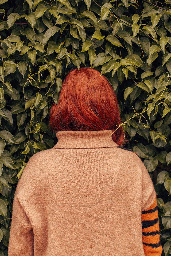 woman with red hair in gray, orange, and black sweater facing on green plant