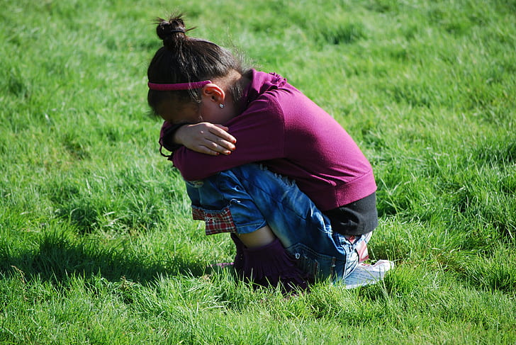 girl wearing maroon long-sleeved top and blue jeans sitting on green grasses during daytime