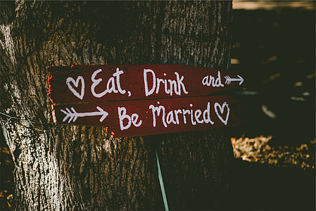 two brown-and-white eat, drink, and be married-printed wooden board signage leaning on tree trunk