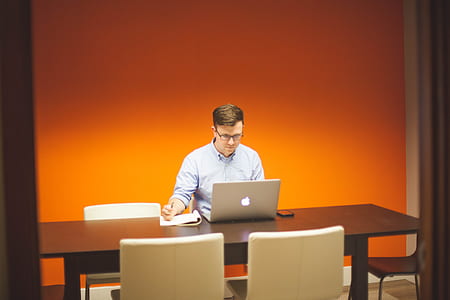 man in blue dress shirt wearing eyeglasses sitting on chair while looking at the silver iMac and holding pen