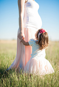 girl in white and pink puff sleeve dress near pregnant mother standing on green grass during daytime