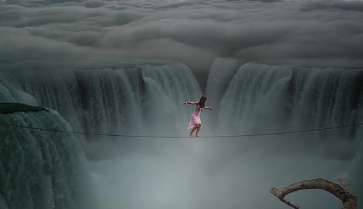 photograph of woman about to cross on rope above waterfalls