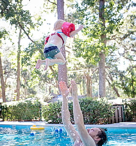 Photography of Man on Swimming Pool Tossing Toddler Above Pool