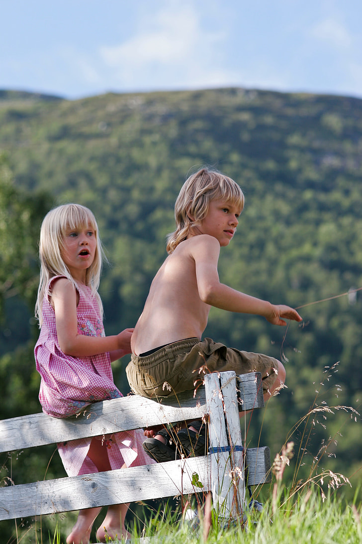 boy and girl sitting on white wooden fence near mountains