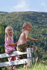 boy and girl sitting on white wooden fence near mountains