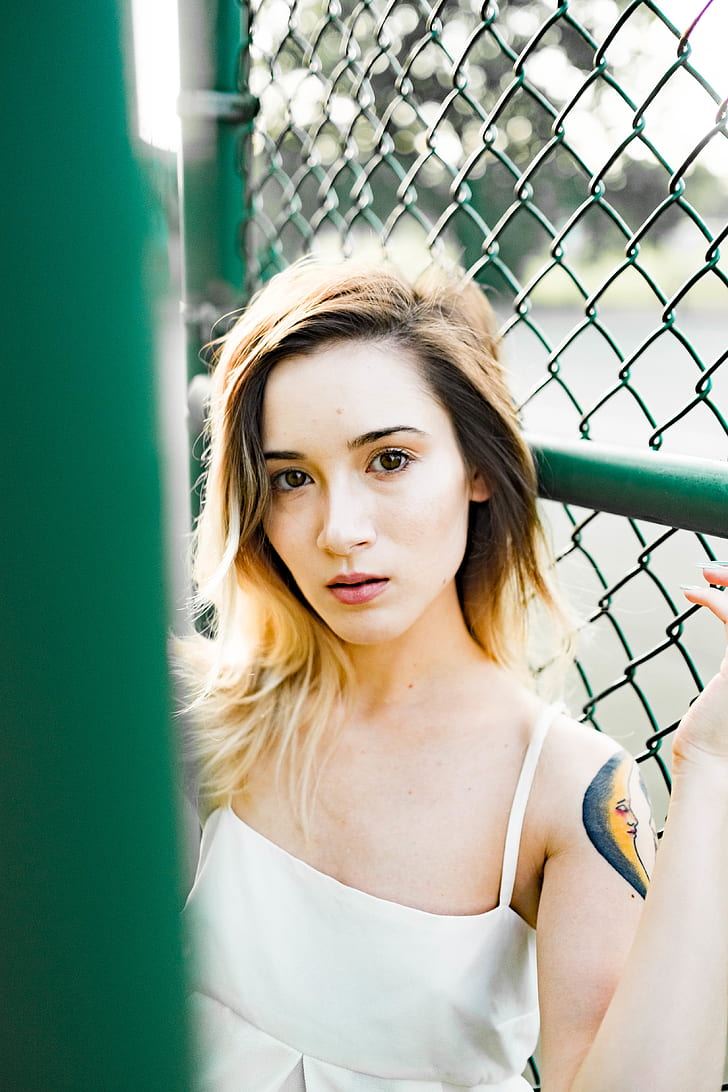 woman wears white sleeveless top leaning on green fence