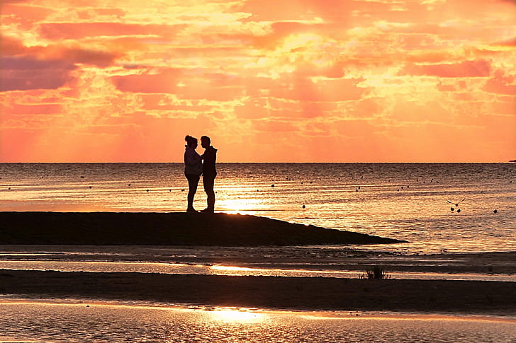 silhouette of man and woman on shore under orange sunset
