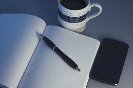 clear black-lined white notebook near pen and coffee mug