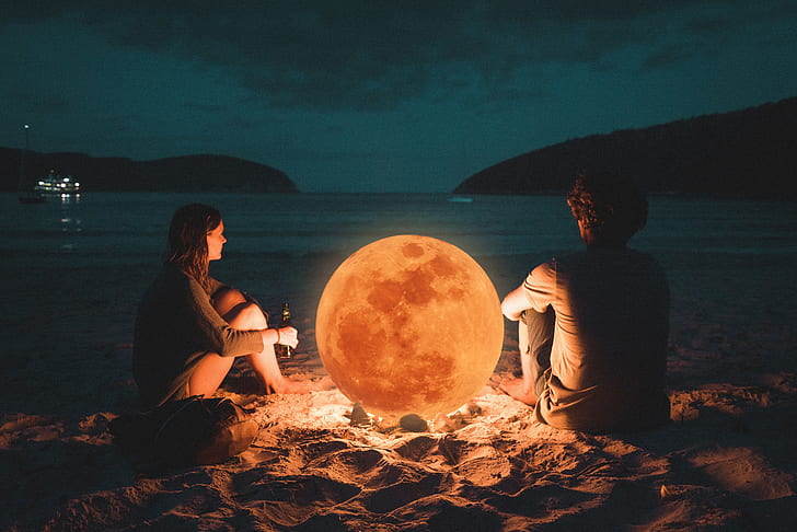 man and woman sitting on sand near sea shore during night time