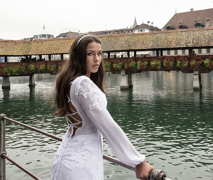 woman wearing white applique long-sleeved strappy-back dress holding gray metal railings near body of water
