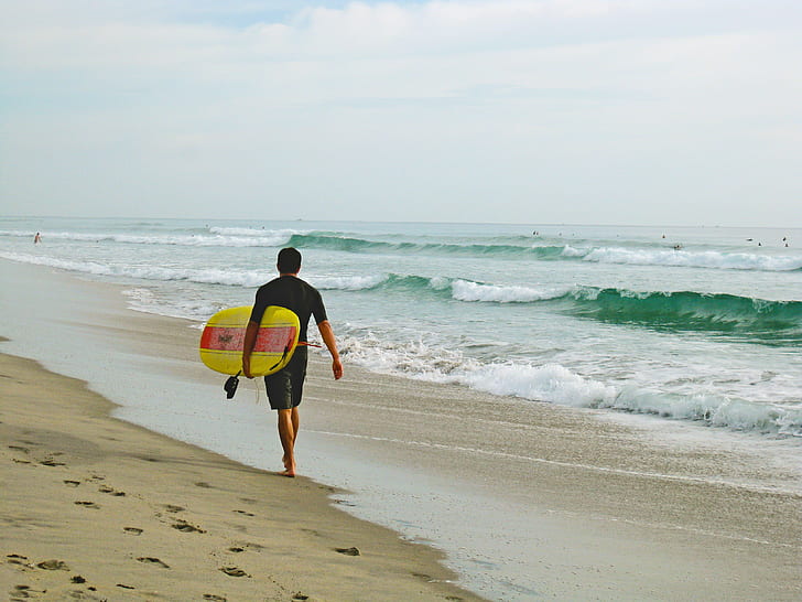 man in black shirt and shorts carrying a yellow surfboard on beach