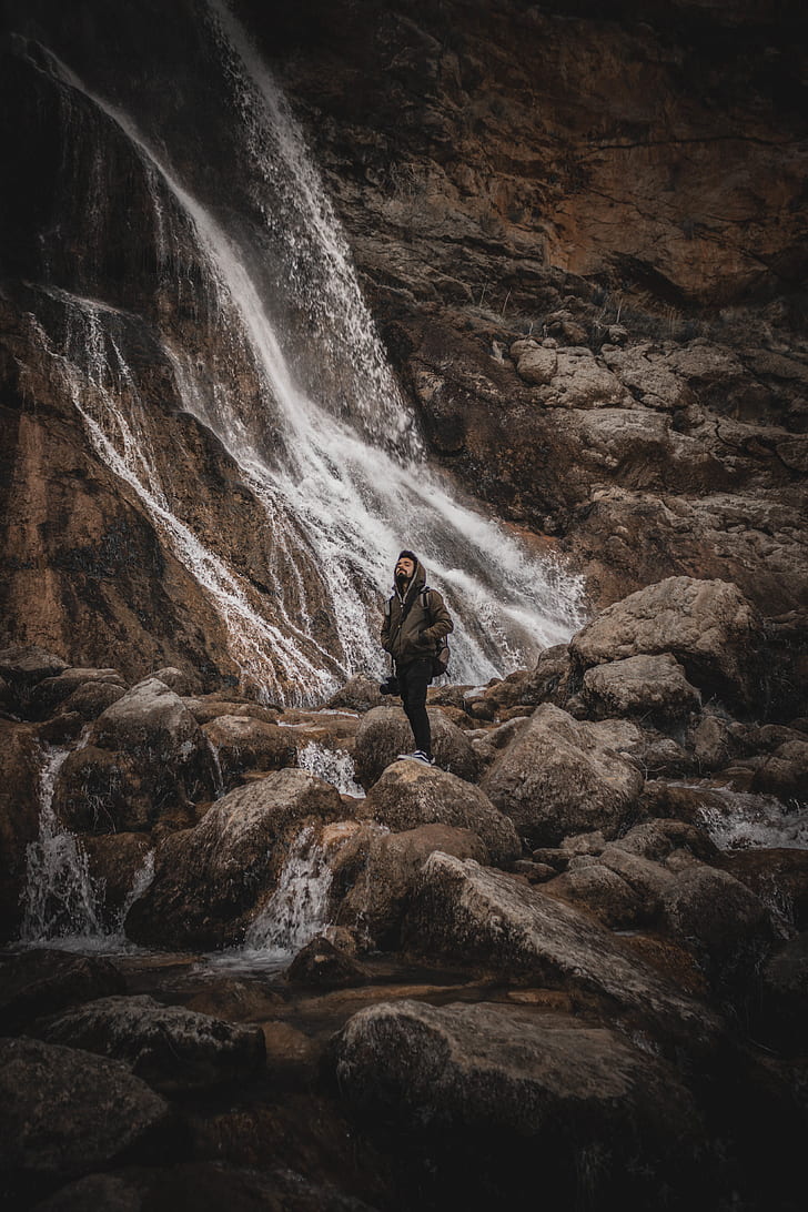 photo of man wearing brown jacket near waterfalls
