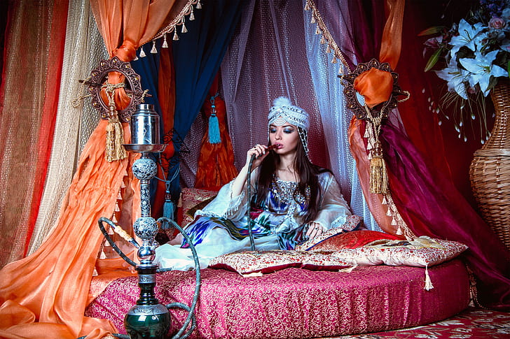 woman wearing blue long-sleeved dress and pagri hat sitting on bed smoking hookah