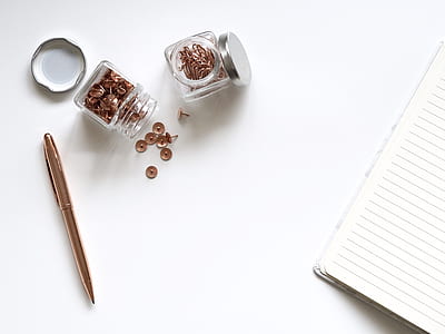 two jars of thumbtacks beside lined notebook and gold retractable pen