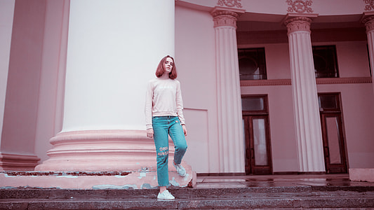 woman wearing pink long-sleeved shirt and blue pants leaning on white pillar during daytime