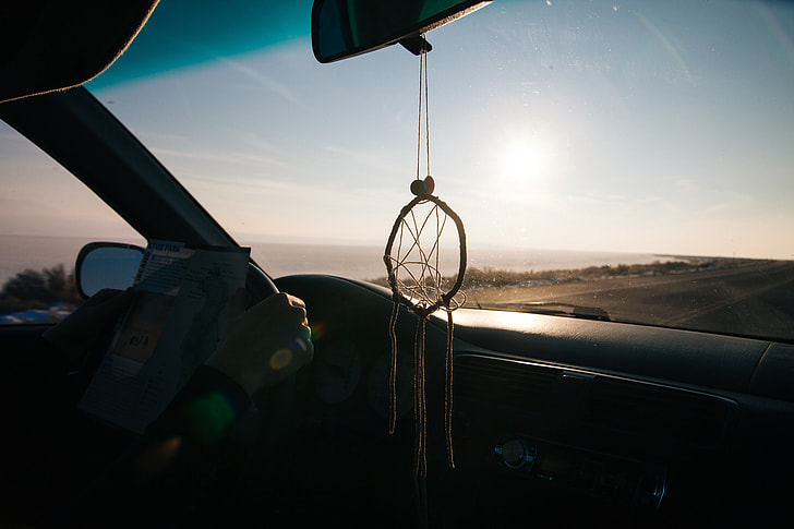 dreamcatcher hanged from rear view mirror inside car driving on road along shore during golden hour